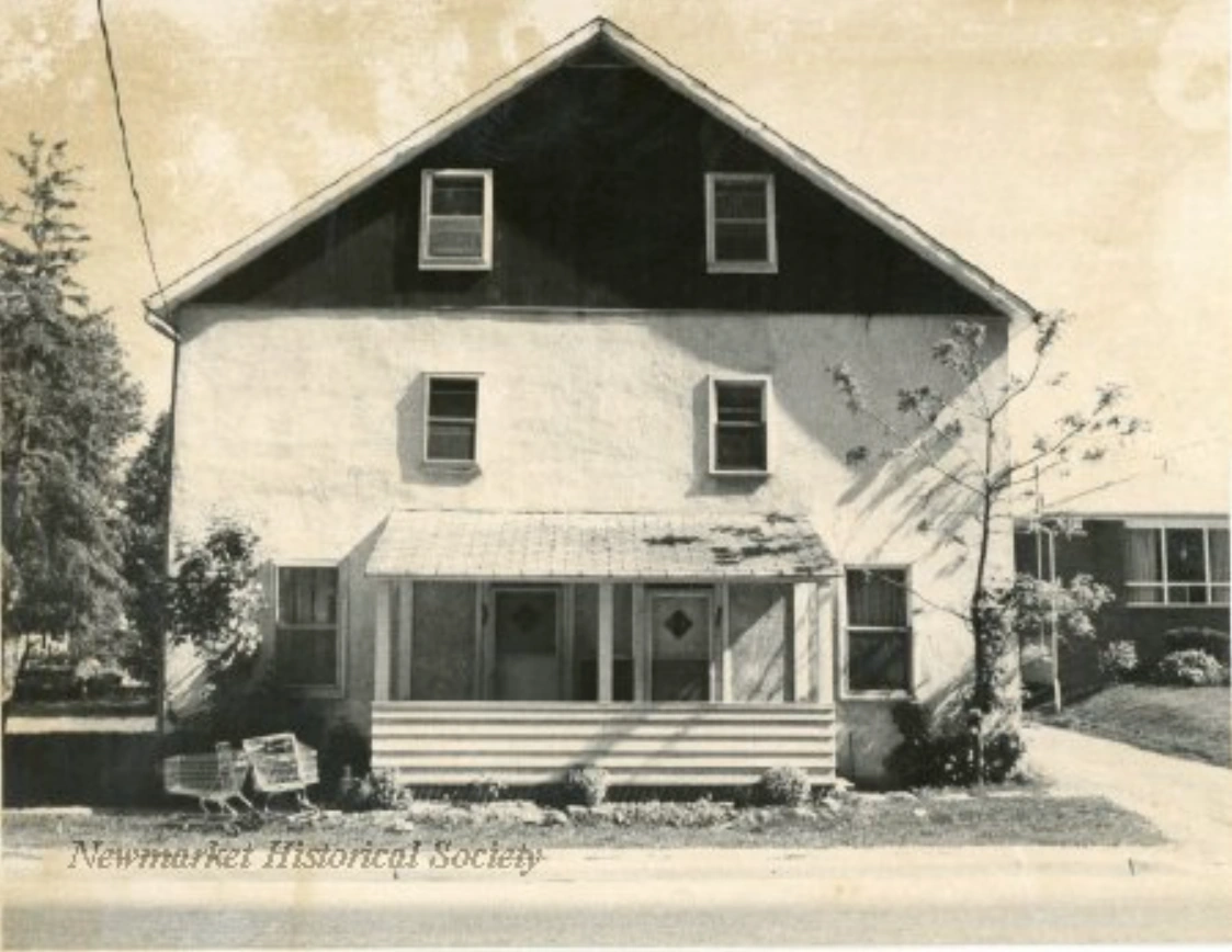 The house around 1976, with a light-coloured wall surface, darker gable, covered entrance and two shopping carts at the roadside